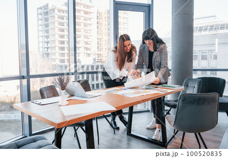 Standing and talking. Two women in formal clothes is indoors in the modern office works together 100520835