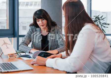 Sitting by the table. Two women in formal clothes is indoors in the modern office works together 100520841