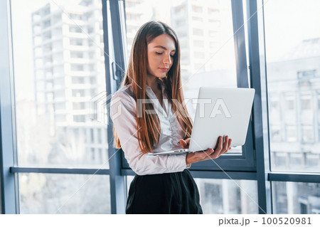Standing with laptop. Young woman in white formal clothes is indoors in the modern office 100520981