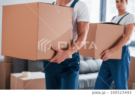 With boxes in hands. Two young movers in blue uniform working indoors in the room 100523493