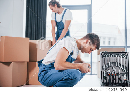 Uses tools from case. Two young movers in blue uniform working indoors in the room Uses tools from case. Two young movers in blue uniform working indoors in the room 100524340