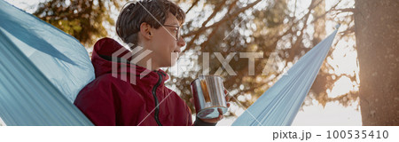 Young woman in glasses sitting in hammock with thermos in forest and enjoying morning. Young woman in glasses sitting in hammock with thermos in forest and enjoying morning. 100535410