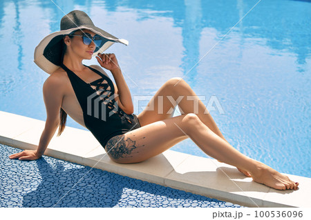 Portrait of a female having rest and posing near a swimming pool. Dressed in a black swimsuit, hat and sunglasses. Portrait of a female having rest and posing near a swimming pool. Dressed in a black swimsuit, hat and sunglasses. 100536096