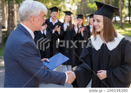 The teacher shakes hands with the student and presents the diploma outdoors. A group of university graduates. 100536260