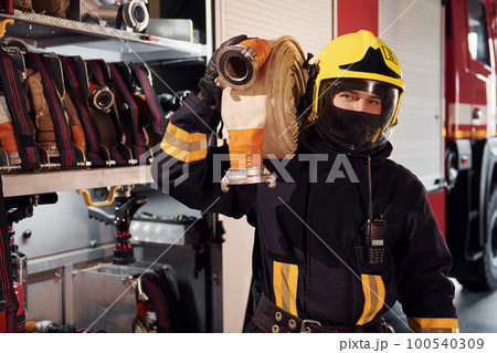 Holds hose. Male firefighter in protective uniform standing near truck 100540309