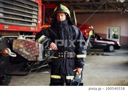 Holds axe in hands. Male firefighter in protective uniform standing near truck Holds axe in hands. Male firefighter in protective uniform standing near truck 100540558