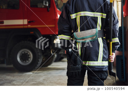 Holds axe in hands. Male firefighter in protective uniform standing near truck Holds axe in hands. Male firefighter in protective uniform standing near truck 100540594