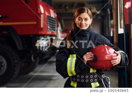 Holds red hat in hands. Female firefighter in protective uniform standing near truck 100540665