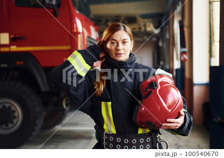 Holds red hat in hands. Female firefighter in protective uniform standing near truck Holds red hat in hands. Female firefighter in protective uniform standing near truck 100540670