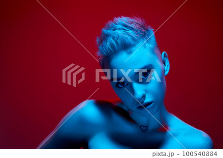 Uniqueness. Beautiful young girl with short blonde hair posing with bare shoulders against red studio background in blue neon light. Cyberpunk style 100540884