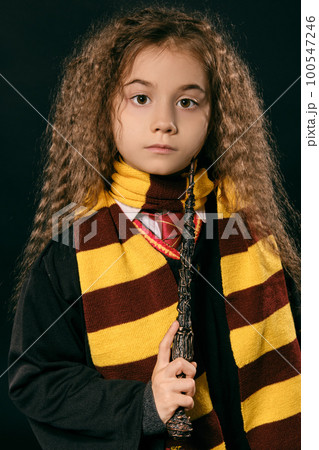 Portrait of a little witch girl with long brown hair dressed in dark coat, holding magic wand in her hand, posing on black studio background. Portrait of a little witch girl with long brown hair dressed in dark coat, holding magic wand in her hand, posing on black studio background. 100547246