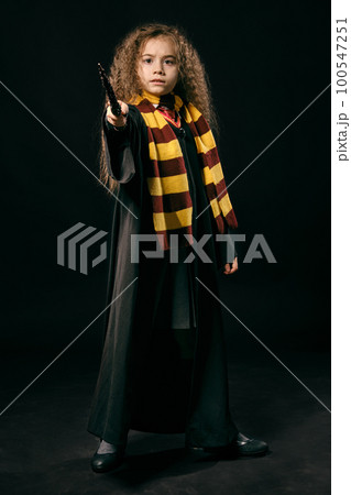 Portrait of a little witch girl with long brown hair dressed in dark coat, holding magic wand in her hand, posing on black studio background. 100547251