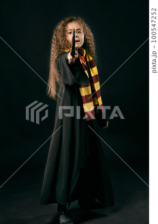 Portrait of a little witch girl with long brown hair dressed in dark coat, holding magic wand in her hand, posing on black studio background. 100547252