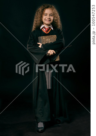 Portrait of a little witch girl with long brown hair dressed in dark coat, holding magic wand in her hand, posing on black studio background. Portrait of a little witch girl with long brown hair dressed in dark coat, holding magic wand in her hand, posing on black studio background. 100547253