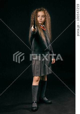 Portrait of a little witch girl with long brown hair dressed in dark clothes, holding magic wand in her hand, posing on black studio background. 100547259