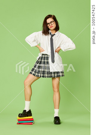 Exam session. Young beautiful woman, student in checkered skirt and white shirt posing with books against green studio background 100547425