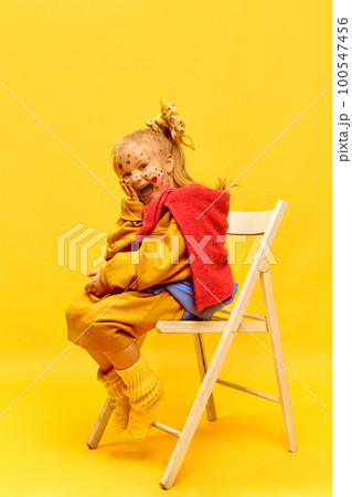 Photo of sweet little girl wearing bow, sitting on chair and laughing at camera over yellow background. Full-length Photo of sweet little girl wearing bow, sitting on chair and laughing at camera over yellow background. Full-length 100547456