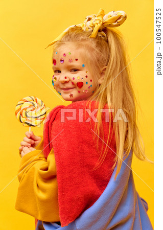 Profile photo of little girl holding lollipop and smiling over yellow background. Concept of fashion, ad, children's products, child model, clothing and accessories Profile photo of little girl holding lollipop and smiling over yellow background. Concept of fashion, ad, children's products, child model, clothing and accessories 100547525