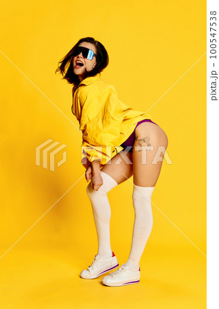 Energy and positivity. Beautiful young brunette woman in stylish sportswear and sunglasses posing against bright yellow studio background. Concept of sport 100547538