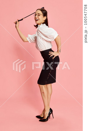Playful punishment. Portrait of young smiling beautiful woman in formal wear posing with whip against pink studio background. Playful punishment. Portrait of young smiling beautiful woman in formal wear posing with whip against pink studio background. 100547708