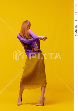 Faceless portrait of slim woman with long straight red hair wearing retro fashion outfit in action over yellow studio background. Impersonal emotions, body language 100547828