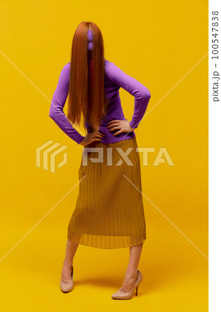 Faceless portrait of slim woman with long straight red hair wearing retro fashion outfit in action over yellow studio background. Impersonal emotions, body language 100547838