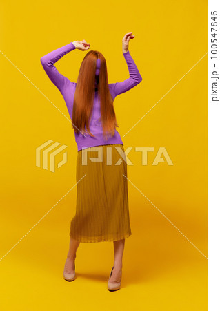 Faceless portrait of slim woman with long straight red hair wearing retro fashion outfit in action over yellow studio background. Impersonal emotions, body language 100547846