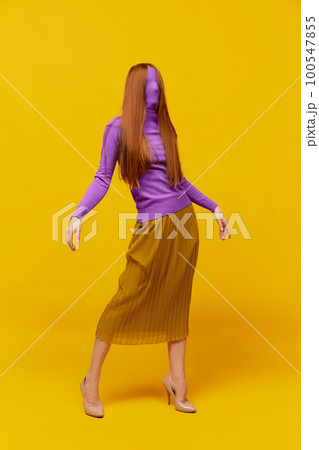 Faceless portrait of slim woman with long straight red hair wearing retro fashion outfit in action over yellow studio background. Impersonal emotions, body language 100547855