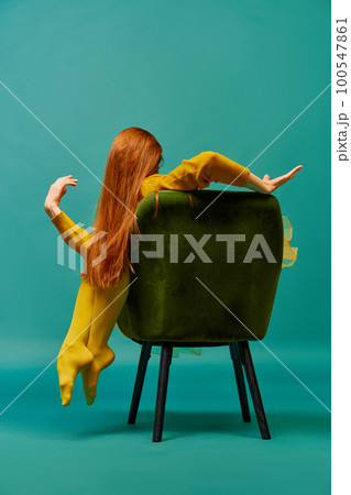 Creative portrait of unknown redhead girl with long straight silky hair posing on armchair, sitting in strange poses over cyan color background. Impersonal emotions, body language 100547861