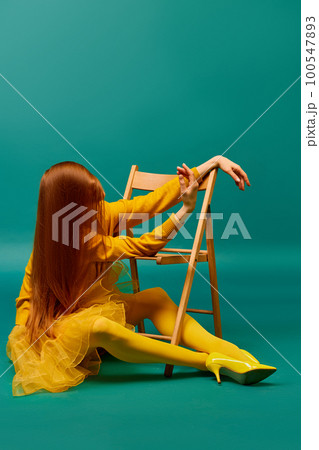 Creative portrait of unknown redhead girl with long straight silky hair posing on armchair, sitting in strange poses over cyan color background. Impersonal emotions, body language 100547893