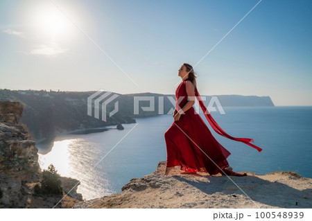 A woman in a red flying dress fluttering in the wind, against the backdrop of the sea. 100548939