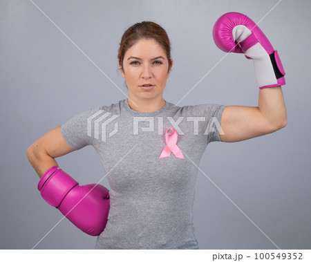 A woman with a pink ribbon on her chest in pink boxing gloves on a gray background. Victory over breast cancer.  100549352