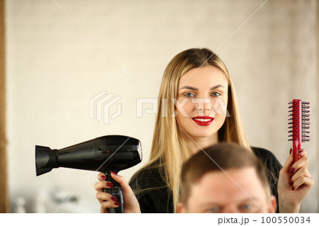 Woman hairdresser is holding hair dryer and comb man is sitting in chair Woman hairdresser is holding hair dryer and comb man is sitting in chair 100550034