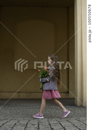 Adorable 9 years old girl with long curly hair walking through central square of the town Olesnica, Poland. Beautiful girl walking with bouquet of tulip flowers through spring European city. 100550678