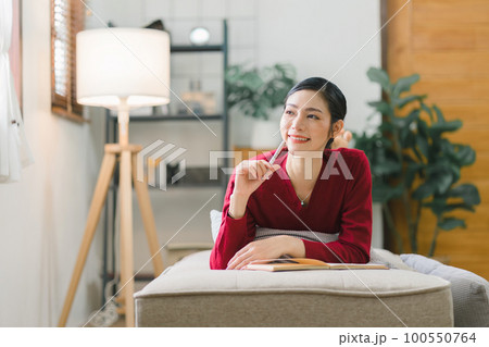 A beautiful young Asian woman is lying on a sofa, writing in her diary with a pen and notebook in her hands. She is a smiling brunette lady. 100550764