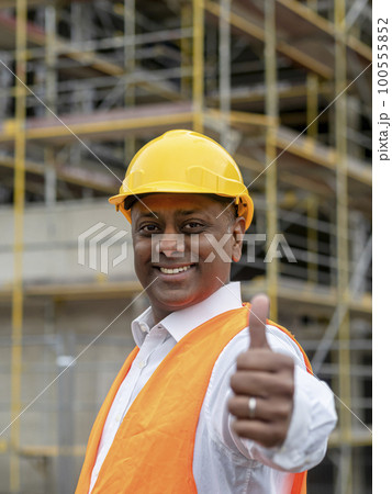 Smiling Indian construction worker wearing safety helmet and vest posing on construction site showing thumbs up gesture Smiling Indian construction worker wearing safety helmet and vest posing on construction site showing thumbs up gesture 100555852