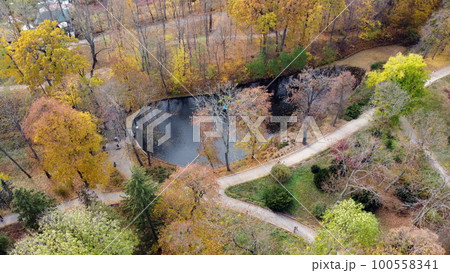Flying over the autumn park. Many trees with yellow green and fallen leaves, lakes, people walking along dirt paths in park on autumn day. Top view. Aerial drone view. Beautiful natural background Flying over the autumn park. Many trees with yellow green and fallen leaves, lakes, people walking along dirt paths in park on autumn day. Top view. Aerial drone view. Beautiful natural background 100558341