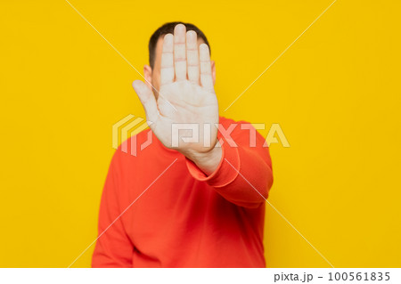Hispanic man putting hand in front to stop camera, rejecting photos or images against yellow colored wall. Rejection and privacy concept. 100561835