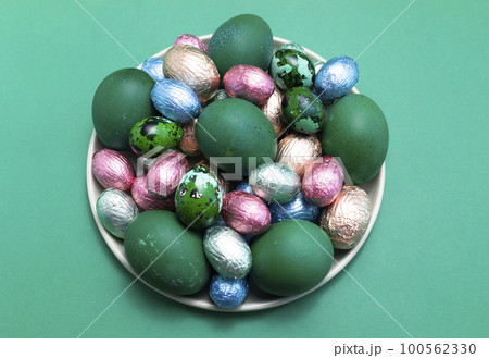 Delicious Easter food, treat. Colored chicken and quail eggs, chocolate eggs, sweets wrapped in bright foil on plate, flatly top view. Close up, horizontal. Christian holiday tradition. Confectionary Delicious Easter food, treat. Colored chicken and quail eggs, chocolate eggs, sweets wrapped in bright foil on plate, flatly top view. Close up, horizontal. Christian holiday tradition. Confectionary 100562330