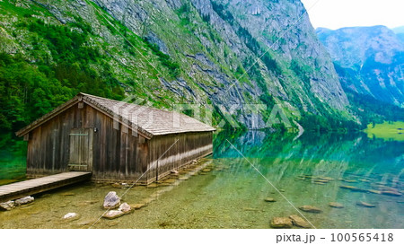 Konigssee, Germany - June 16, 2018: side view of lakeside cabin or traditional wooden boat house in Obersee on the left with a peaceful and clear lake on a sunny summer day Konigssee, Germany - June 16, 2018: side view of lakeside cabin or traditional wooden boat house in Obersee on the left with a peaceful and clear lake on a sunny summer day 100565418