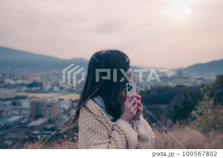 朝日を浴び街をバックに佇むスマホを持った女性の写真 100567802
