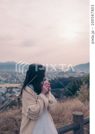 朝日を浴び街をバックに佇むスマホを持った女性の写真 100567803