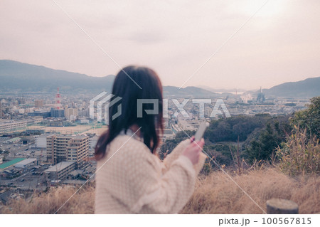 朝日を浴び街をバックに佇むスマホを持った女性の写真 朝日を浴び街をバックに佇むスマホを持った女性の写真 100567815