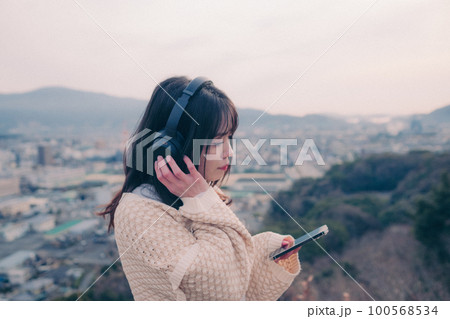 朝日を浴び街をバックに音楽を聴く女性の写真 100568534