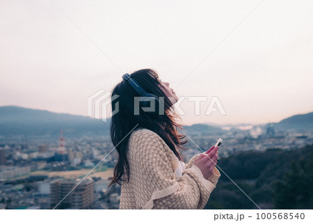 朝日を浴び街をバックに音楽を聴く女性の写真 100568540