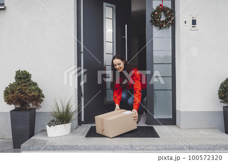 Young woman in red Christmas sweater picking up a cardboard box mailman left out at the front door 100572320