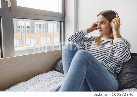 Blonde woman college student relaxing on her bed listening to some music while looking out the window Blonde woman college student relaxing on her bed listening to some music while looking out the window 100572346