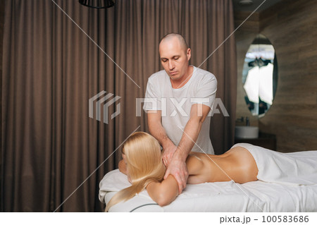 Medium shot of male masseur massaging shoulder of young woman lying on table in spa salon. Female patient getting remedial body and shoulder blade massage easing pain and relaxing muscles. 100583686
