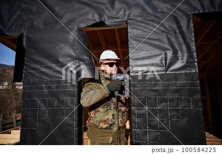 Male builder installing waterproof membrane on the wall of future cottage. Man worker building wooden frame house. Carpentry and construction concept. Male builder installing waterproof membrane on the wall of future cottage. Man worker building wooden frame house. Carpentry and construction concept. 100584225