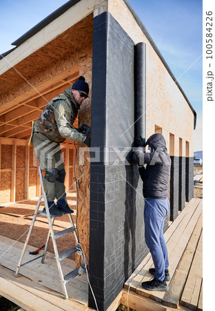 Male builders installing waterproof membrane on the wall of future cottage. Men workers building wooden frame house in the Scandinavian style barnhouse. Carpentry and construction concept. 100584226